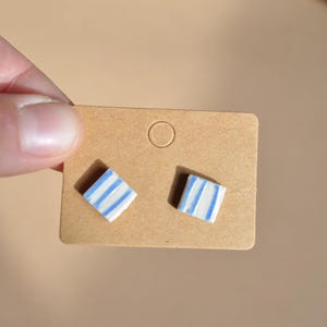 Puede incluir: Un par de pendientes cuadrados con un diseño de rayas azules y blancas. Los pendientes se muestran en una pequeña tarjeta de pendientes marrón. Los pendientes son pequeños y parecen hechos a mano.