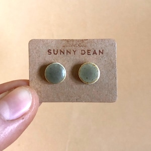 May include: A pair of round, sage green stud earrings on a brown card. The card is labeled "SUNNY DEAN" in a simple font. The earrings are held by a hand.