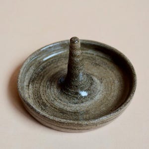 May include: A handmade ceramic ring holder in a muted brown color. The ring holder has a circular base with a raised, conical center for holding rings. The surface has a textured, earthy appearance, with visible spiral patterns.