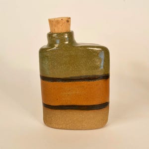 May include: A rectangular ceramic bottle with a cork stopper. The bottle features horizontal bands of olive green, orange, and tan, separated by thin black lines. The cork is a light brown color.