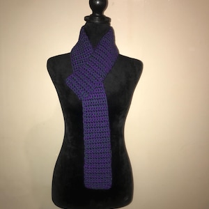 Puede incluir: Bufanda de ganchillo morada y azul oscuro hecha a mano, exhibida en un maniquí negro. La bufanda es de color liso con un aspecto texturizado, probablemente hecha de hilo. La bufanda está drapeada alrededor del cuello del maniquí.