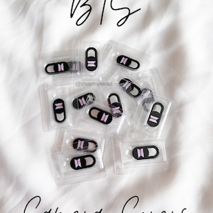 May include: A group of black camera covers with a pink BTS logo. The covers are packaged in clear plastic. The text "BTS Camera Covers" is written below the covers.