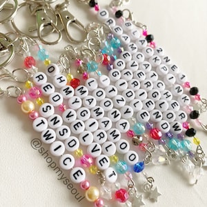 May include: A set of 10 colorful beaded keychains with different K-pop group names spelled out on white beads. The keychains feature a variety of colorful beads, including clear, pink, red, blue, and yellow. Each keychain has a silver key ring and a charm at the end, either a silver star or a silver heart.