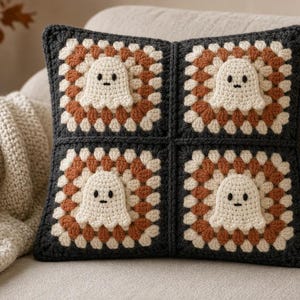 May include: A square crochet pillow featuring four granny square panels joined together. Each panel is dark charcoal gray with a rust orange and cream center, and each features a small, cream-colored ghost applique with black eyes and a mouth.