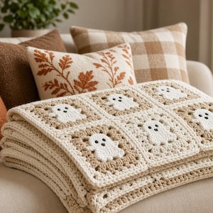 Könnte beinhalten: Eine gehäkelte Decke mit einem sich wiederholenden Muster aus weißen Geistermotiven, die in beigen und cremefarbenen Granny Squares zentriert sind. Die Decke ist gefaltet und zeigt eine strukturierte Garnkonstruktion mit sichtbaren Maschen.