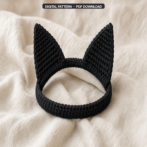 May include: A black crocheted headband featuring two pointed cat ears. The item is a digital pattern and PDF download. The headband is constructed with a dense, textured stitch pattern and forms a circular shape with upright triangular ears.