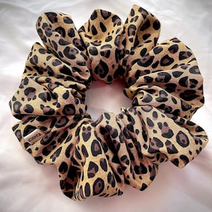 May include: A large scrunchie with a leopard print pattern. The scrunchie is made of a soft fabric with a tan background and dark brown spots. The scrunchie is gathered and has a label that reads "VERRUSA".