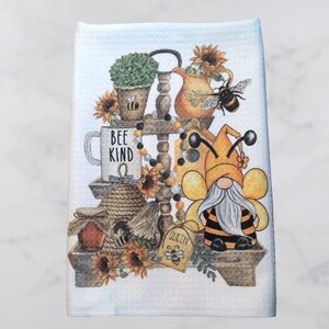 May include: A white kitchen towel with a bee-themed design. The design features a gnome wearing a bee costume, a beehive, a mug that says "Bee Kind", and sunflowers.