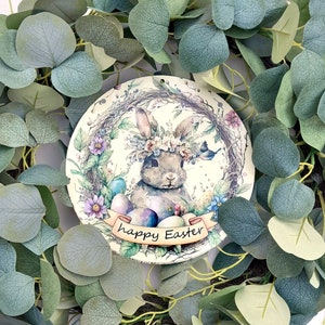 May include: A round wooden ornament with a watercolor illustration of a bunny wearing a flower crown and holding Easter eggs. The bunny is surrounded by flowers and greenery. The ornament has the text "happy Easter" written on it.