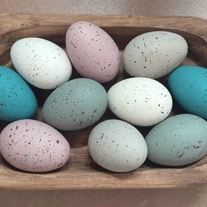 May include: A wooden bowl filled with eleven speckled decorative eggs in various pastel colors: white, pink, teal, and blue. The eggs are oval-shaped and have a speckled pattern. The bowl is rectangular with rounded edges.