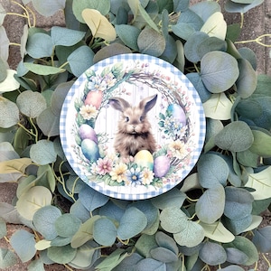 May include: A blue and white checkered coaster with a floral design and a brown bunny rabbit in the center. The bunny is surrounded by colorful eggs and flowers.