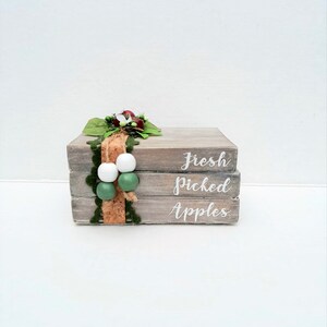 May include: A stack of three wooden blocks with the words "Fresh Picked Apples" painted in white. The blocks are decorated with a green and white garland, a brown ribbon, and a cork.