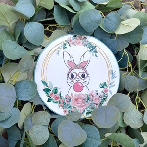 May include: A round decorative plate featuring a cartoon bunny with heart-shaped glasses blowing a bubblegum bubble. The bunny is surrounded by pink roses and green leaves, with a gold border. The plate is set against a backdrop of green foliage.