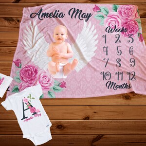 Puede incluir: Manta rosa para bebé con diseño floral y alas de ángel blancas. La manta tiene una tabla de crecimiento con números para semanas y meses. El texto "Amelia May" está impreso en la manta.