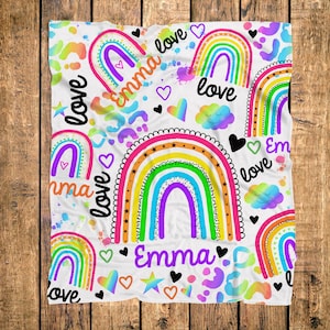 May include: A white blanket with a colorful rainbow pattern, hearts, stars, and the name "Emma" in a playful font. The word "love" is also featured. The design includes a mix of bright colors and a whimsical aesthetic.