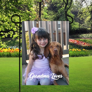 May include: A young girl with a purple flower in her hair sits on a wooden deck with a brown dog. The girl is wearing a purple dress and the dog is looking at the camera. The text "Grandma Loves" is written in white cursive on the bottom of the image.