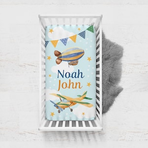 May include: A blue and yellow baby crib sheet with a white background. The sheet features a yellow airplane, a blue and yellow airship, and white clouds. The sheet also has the name "Noah John" printed on it.