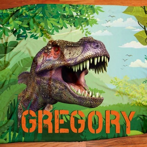 May include: A green and blue blanket with a dinosaur and jungle leaves. The dinosaur is green, purple, and brown with a large open mouth. The text "GREGORY" is printed in orange on the blanket.
