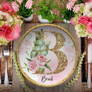 May include: A decorative plate with a gold glitter initial "B" and the name "Brook" in script. The plate features pink floral and greenery accents, with a gold rim. The plate is set on a wooden table with silverware and floral arrangements.