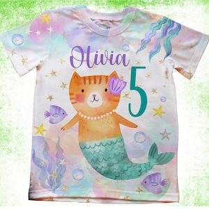 May include: White t-shirt with a colourful mermaid cat design. The shirt features the name "Olivia" and the number "5". The design includes a cat with a mermaid tail, fish, bubbles, and stars.