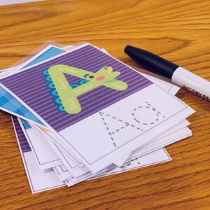 K&ouml;nnte beinhalten: Ein Stapel violetter und wei&szlig;er Karteikarten mit einem gr&uuml;nen Cartoon-Dinosaurier-Buchstaben A und einer gestrichelten Linie zum Nachzeichnen. Ein Trockenl&ouml;schmarker liegt neben dem Stapel.