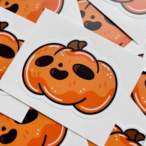 May include: Orange pumpkin sticker with a smiling face and a brown stem. The sticker has a white border and is surrounded by other similar stickers. The pumpkin design is cartoonish and suitable for Halloween or fall-themed crafts.