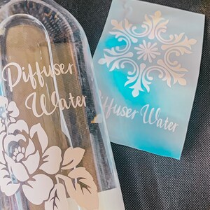 May include: A clear glass bottle with a metal lid and a floral design. The bottle is labeled "Diffuser Water". A separate sheet of paper with a decorative design and the text "Diffuser Water" is also visible.