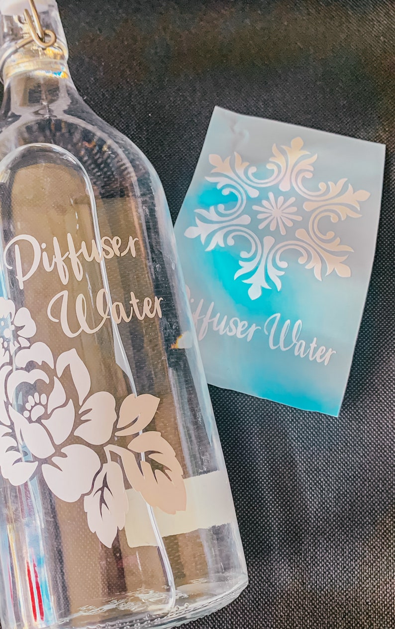 May include: A clear glass bottle with a metal lid and a floral design. The bottle is labeled "Diffuser Water". A separate sheet of paper with a decorative design and the text "Diffuser Water" is also visible.