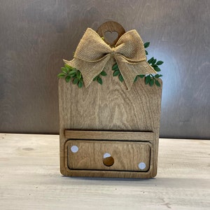 May include: A wooden cutting board with a heart-shaped cutout and a burlap bow. The board has a small shelf and two white dots on the bottom.