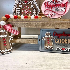 K&ouml;nnte beinhalten: Ein Schild aus Holz f&uuml;r eine Lebkuchen-Kekse-Firma mit einer Lebkuchenmann-Grafik und dem Text "Gingerbread Cookie Company". Das Schild befindet sich auf einem Holzregal mit einem Lebkuchenhaus und einem rosa Schild mit der Aufschrift "Peppermint Wishes".