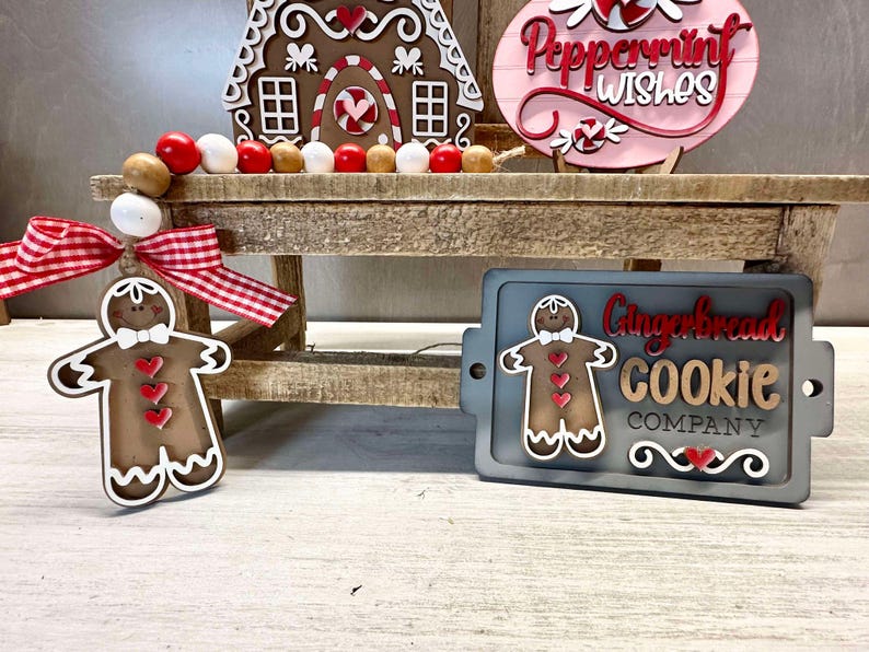 K&ouml;nnte beinhalten: Ein Schild aus Holz f&uuml;r eine Lebkuchen-Kekse-Firma mit einer Lebkuchenmann-Grafik und dem Text "Gingerbread Cookie Company". Das Schild befindet sich auf einem Holzregal mit einem Lebkuchenhaus und einem rosa Schild mit der Aufschrift "Peppermint Wishes".