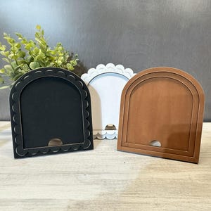 May include: Three wooden arched frames in black, white, and brown. The black frame has a scalloped edge, while the white frame has a scalloped bottom edge. The brown frame has a layered design. Each frame has a small opening at the bottom.
