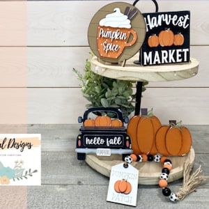 May include: A two-tiered wooden stand decorated with fall-themed items. The top tier features a sign that says "Harvest Market" and a mug with the words "Pumpkin Spice". The bottom tier features a black truck with the words "hello fall pumpkins" and three pumpkins. There is also a tag that says "Pumpkin Patch" with a pumpkin design and a black and white beaded garland.