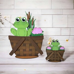May include: Two wooden frog figurines in baskets with pink flowers. The larger frog is sitting in a brown basket with green reeds and a pink flower. The smaller frog is sitting in a brown basket with two pink flowers.