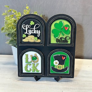 May include: A black wooden St. Patrick's Day decoration with four arched panels. Each panel features a different design: "Lucky" with coins, a shamrock with "Irish", a horseshoe with "Luck", and a leprechaun hat.