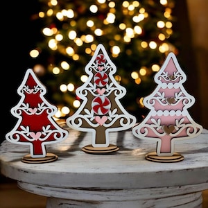 Puede incluir: Tres adornos de &aacute;rbol de Navidad de madera con fondo blanco y recortes intrincados. Los &aacute;rboles est&aacute;n decorados con dise&ntilde;os de bastones de caramelo y corazones rojos, rosas y marrones. Los &aacute;rboles est&aacute;n de pie sobre una mesa de madera blanca.