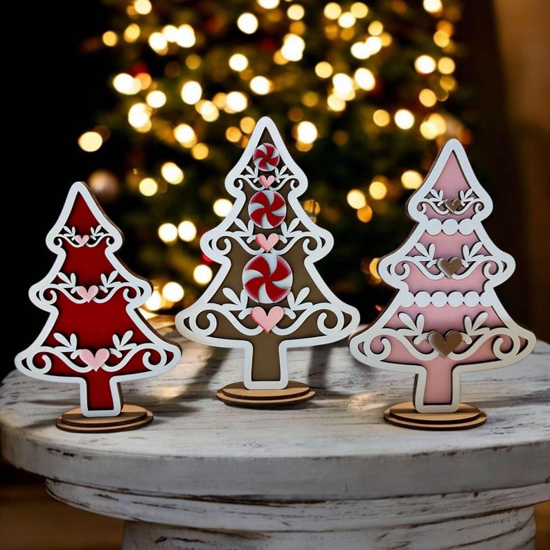 Puede incluir: Tres adornos de &aacute;rbol de Navidad de madera con fondo blanco y recortes intrincados. Los &aacute;rboles est&aacute;n decorados con dise&ntilde;os de bastones de caramelo y corazones rojos, rosas y marrones. Los &aacute;rboles est&aacute;n de pie sobre una mesa de madera blanca.