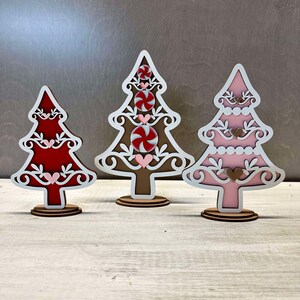 Puede incluir: Tres adornos de &aacute;rbol de Navidad de madera, cada uno con un esquema de color diferente. El primer &aacute;rbol es rojo con detalles blancos, el segundo es marr&oacute;n con detalles blancos y rojos, y el tercero es rosa con detalles blancos. Cada &aacute;rbol tiene un bast&oacute;n de caramelo y corazones.