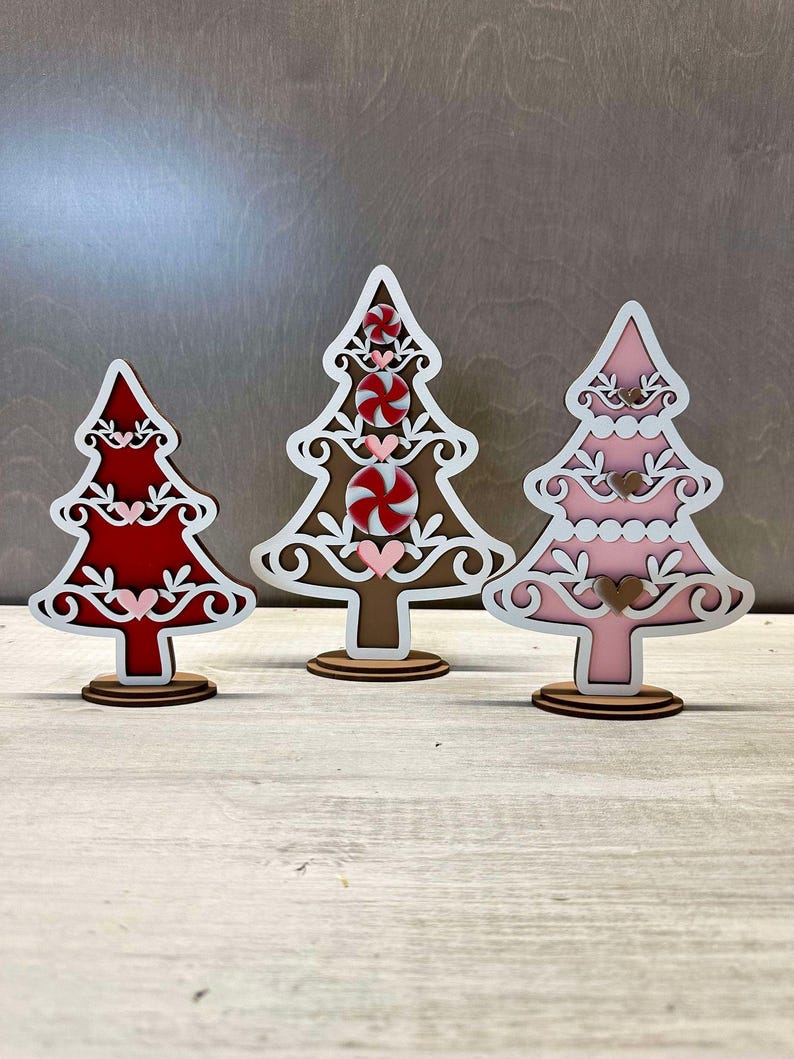 Puede incluir: Tres adornos de &aacute;rbol de Navidad de madera, cada uno con un esquema de color diferente. El primer &aacute;rbol es rojo con detalles blancos, el segundo es marr&oacute;n con detalles blancos y rojos, y el tercero es rosa con detalles blancos. Cada &aacute;rbol tiene un bast&oacute;n de caramelo y corazones.