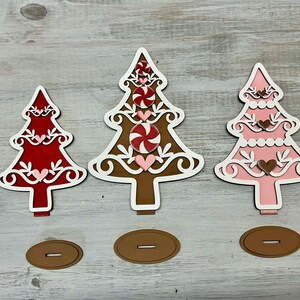 Puede incluir: Tres adornos de &aacute;rbol de Navidad de madera, uno rojo, uno marr&oacute;n y uno rosa. Cada &aacute;rbol est&aacute; decorado con espirales blancas, bastones de caramelo y corazones. Los &aacute;rboles est&aacute;n sobre una superficie de madera.