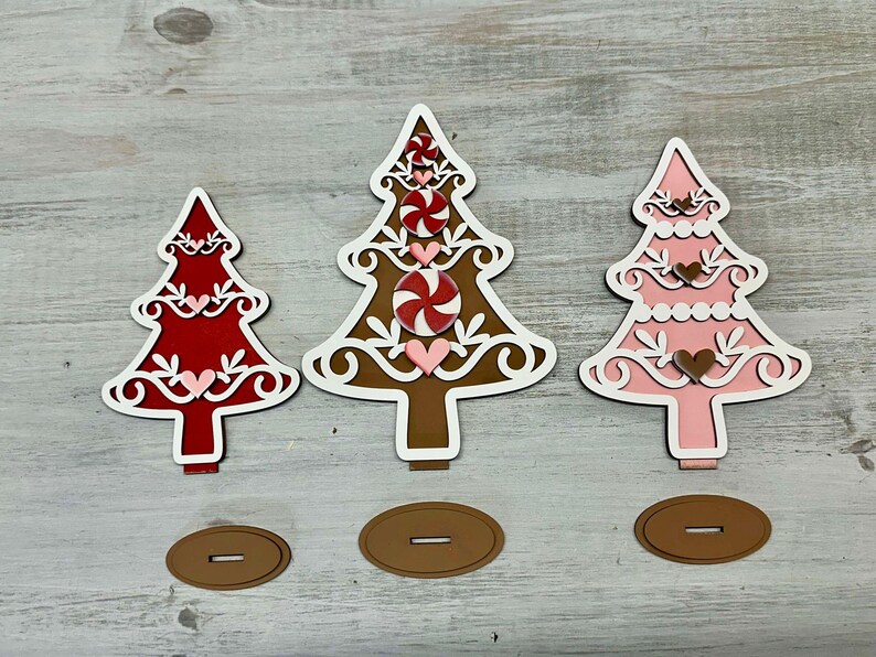 Puede incluir: Tres adornos de &aacute;rbol de Navidad de madera, uno rojo, uno marr&oacute;n y uno rosa. Cada &aacute;rbol est&aacute; decorado con espirales blancas, bastones de caramelo y corazones. Los &aacute;rboles est&aacute;n sobre una superficie de madera.