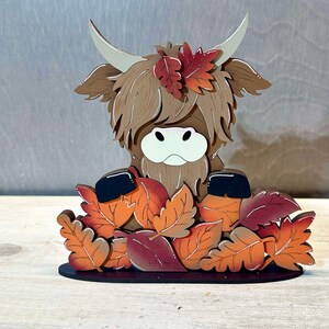 May include: A wooden figurine of a Highland cow with brown fur peeking out from a pile of orange, red, and brown leaves. The cow has a white face and black eyes.