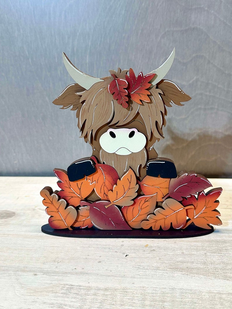May include: A wooden figurine of a Highland cow with brown fur peeking out from a pile of orange, red, and brown leaves. The cow has a white face and black eyes.