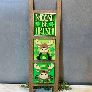 May include: A wooden sign with three panels. The top panel says "Moose Be Irish" with green shamrocks. The middle panel has a brown moose wearing a green shamrock hat and holding a pot of gold. The bottom panel has a brown moose wearing a green leprechaun hat and holding a banner that says "LUCK".