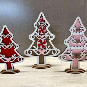 Puede incluir: Tres adornos de &aacute;rbol de Navidad de madera, cada uno con un esquema de color diferente. El primer &aacute;rbol es marr&oacute;n con detalles blancos y dise&ntilde;os de bast&oacute;n de caramelo rojo y blanco. El segundo &aacute;rbol es rojo con detalles blancos y corazones rosas. El tercer &aacute;rbol es rosa con detalles blancos y corazones marrones.