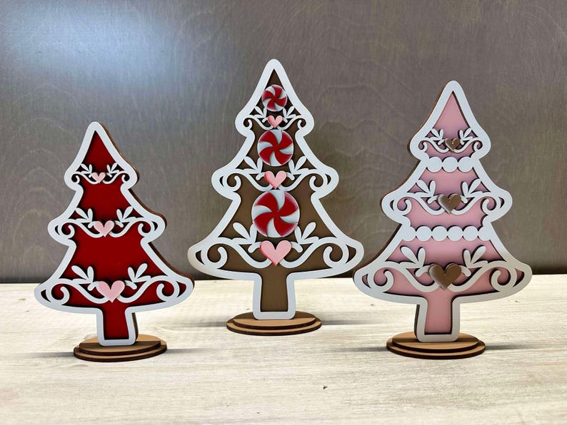 Puede incluir: Tres adornos de &aacute;rbol de Navidad de madera, cada uno con un esquema de color diferente. El primer &aacute;rbol es marr&oacute;n con detalles blancos y dise&ntilde;os de bast&oacute;n de caramelo rojo y blanco. El segundo &aacute;rbol es rojo con detalles blancos y corazones rosas. El tercer &aacute;rbol es rosa con detalles blancos y corazones marrones.