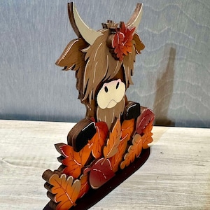 May include: A wooden figurine of a brown Highland cow with a long, shaggy mane. The cow is surrounded by orange and brown leaves and a red apple. The figurine is standing on a brown base.