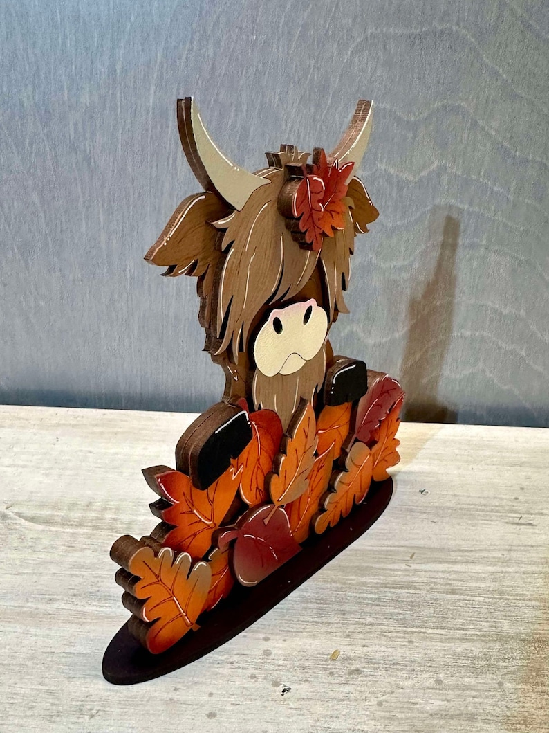 May include: A wooden figurine of a brown Highland cow with a long, shaggy mane. The cow is surrounded by orange and brown leaves and a red apple. The figurine is standing on a brown base.