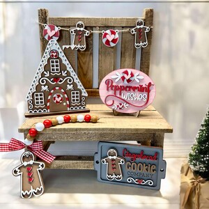 K&ouml;nnte beinhalten: Ein Holz-Lebkuchenhaus, ein Holzschild mit der Aufschrift "Peppermint Wishes", ein Holzschild mit der Aufschrift "Gingerbread Cookie Company" und ein Lebkuchenmann-Anh&auml;nger auf einer Holzbank mit einer rot-wei&szlig; karierten Schleife und einer Perlenkette.