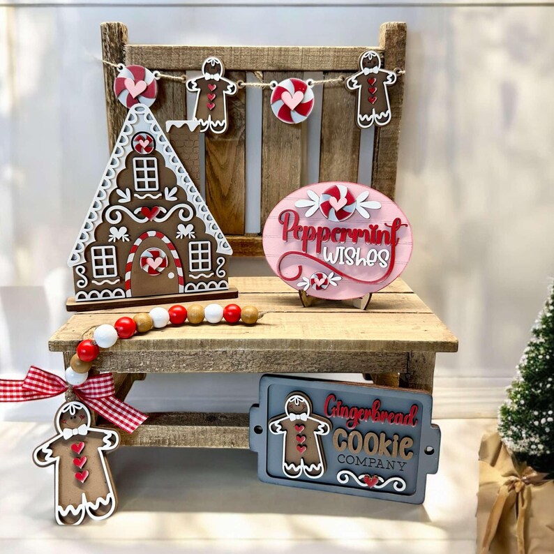 K&ouml;nnte beinhalten: Ein Holz-Lebkuchenhaus, ein Holzschild mit der Aufschrift "Peppermint Wishes", ein Holzschild mit der Aufschrift "Gingerbread Cookie Company" und ein Lebkuchenmann-Anh&auml;nger auf einer Holzbank mit einer rot-wei&szlig; karierten Schleife und einer Perlenkette.